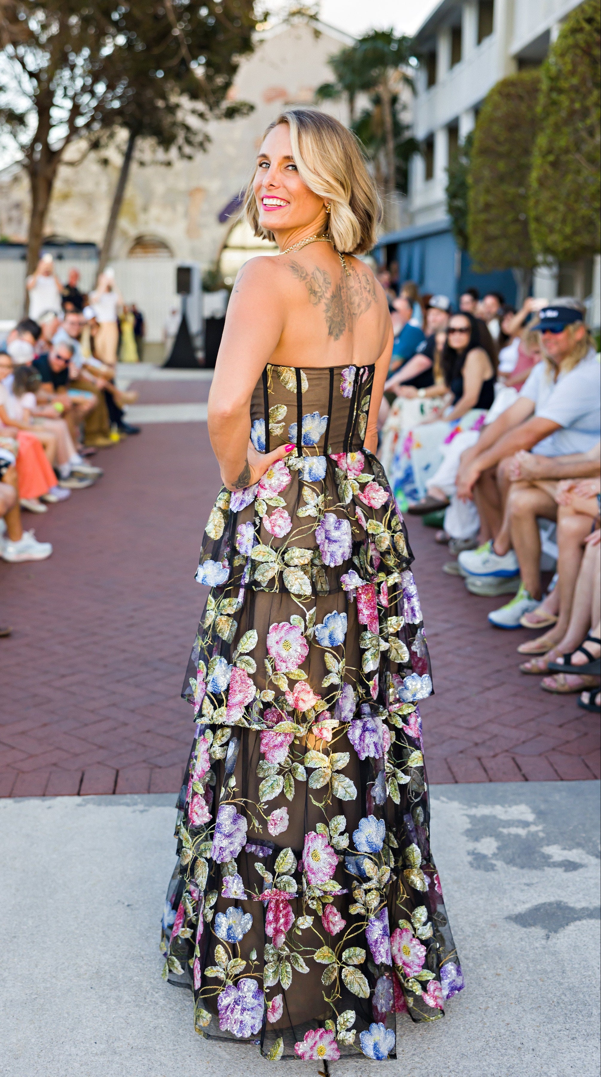 Kirby Myers in a floral dress at an outdoor event with people and buildings in the background